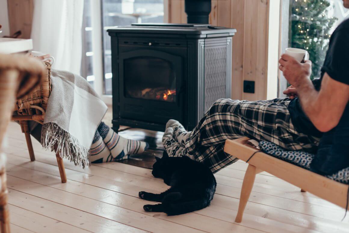 a couple sitting by the fire drinking out of mugs with a black cat on the floor